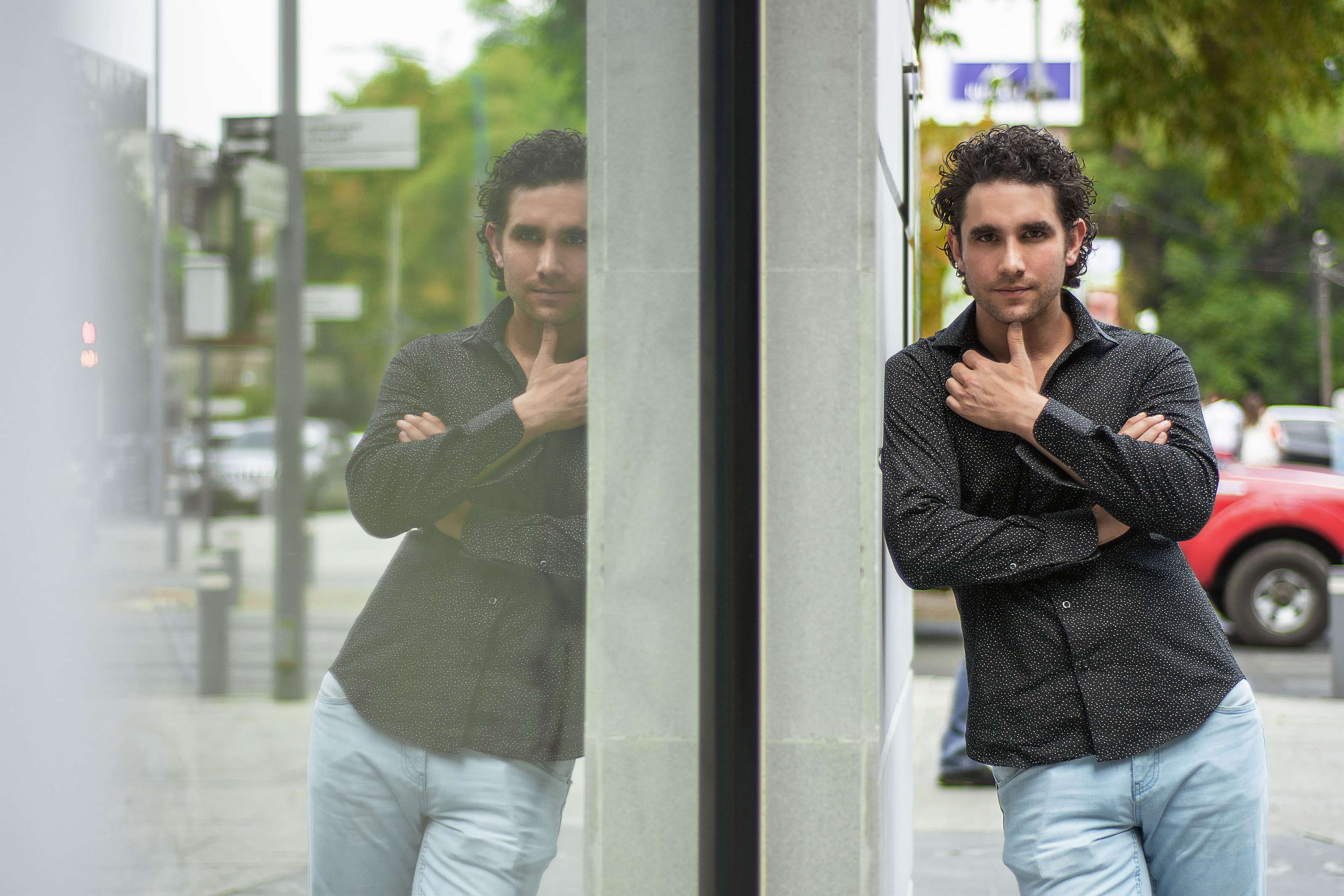 Marco Valdés con camisa negra y jeans, de pie junto a un ventanal